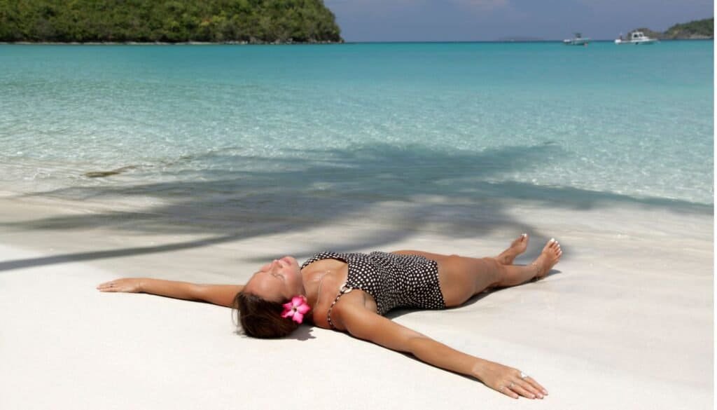 Person in a swimsuit lying on a sandy beach with arms outstretched, next to clear turquoise water. A flower is in their hair, and a shadow of a palm tree is visible.