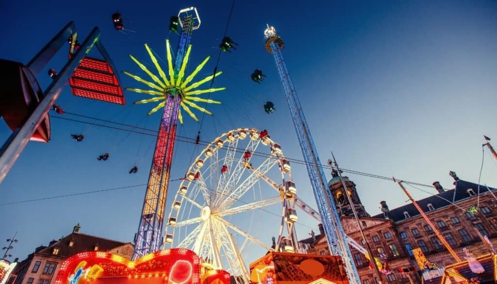A brightly lit carnival scene at dusk featuring a Ferris wheel, a drop tower, and various rides against a clear sky. Buildings are visible in the background.