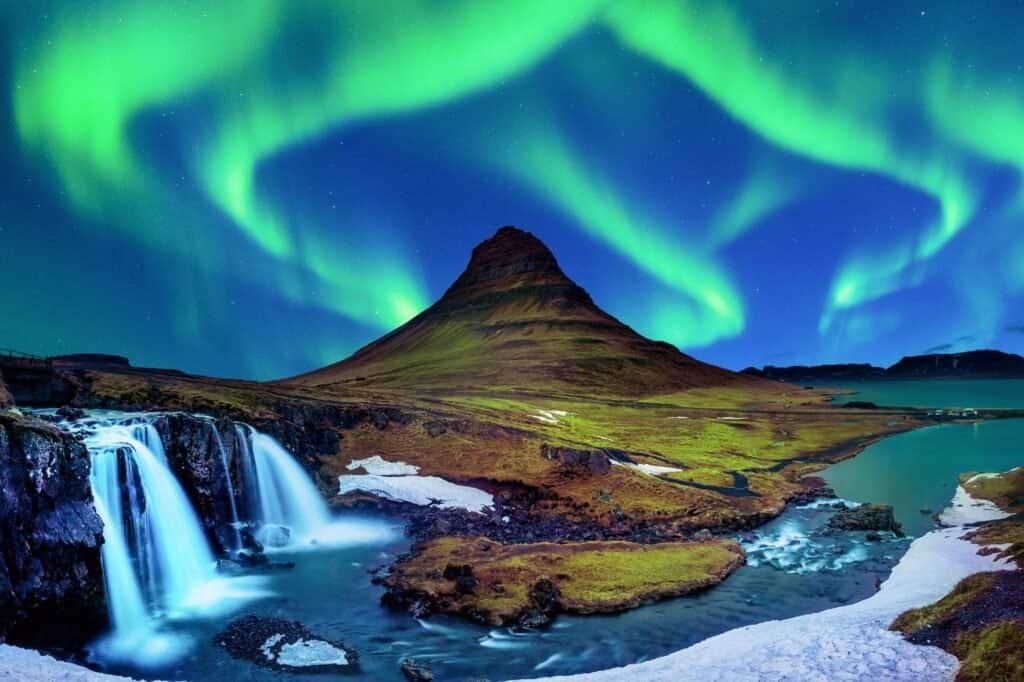 The aurora borealis lights up the night sky above a mountain and waterfall, creating a mesmerizing view in one of the top adventure travel destinations.