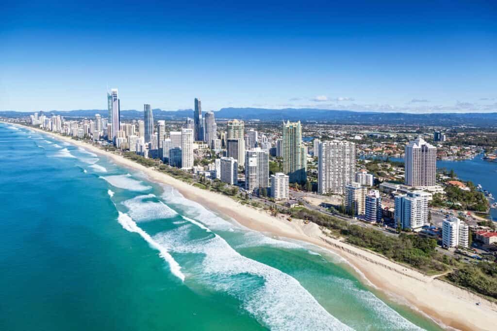 Aerial view of a coastal cityscape, showcasing high-rise buildings along a sandy beach, bordered by turquoise ocean waves under a clear blue sky. It's one of the top adventure travel destinations, offering thrilling activities against a stunning backdrop.