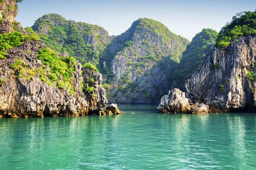 Scenic view of a calm, green bay surrounded by rocky cliffs and lush vegetation under a clear sky, making it one of the top adventure travel destinations.