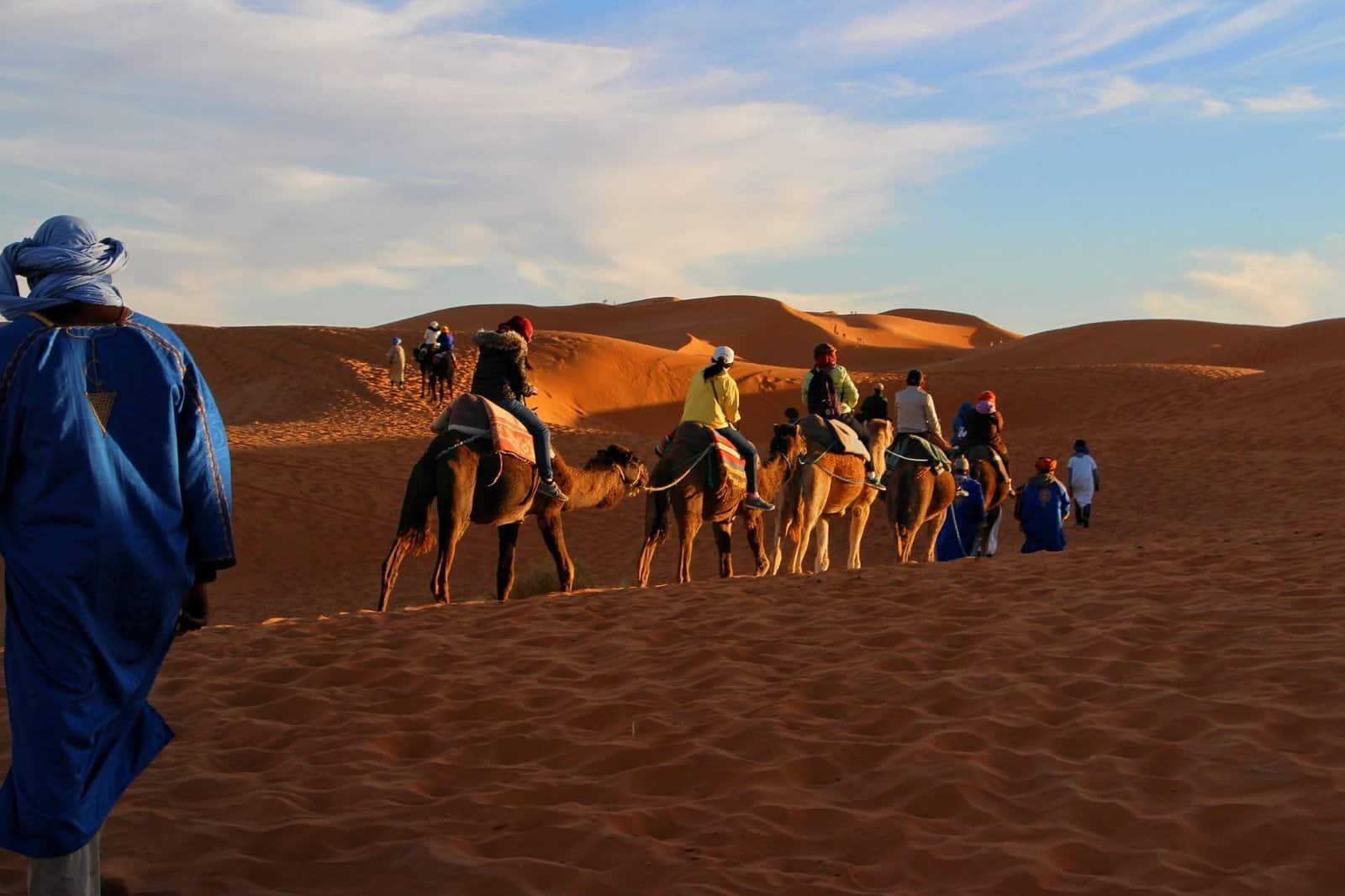 A line of people riding camels traverses through sandy desert dunes under a blue sky, showcasing one of the best adventure travel destinations.