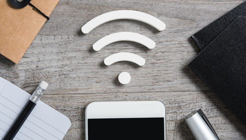 A wooden surface displays a Wi-Fi symbol crafted from white pieces, hinting at travel technology. Below rest notebooks, a smartphone, a pen, and a cardboard box.