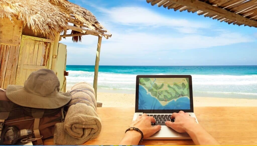 A person is immersed in travel technology, using a laptop with a map on the screen at the beach. Nearby, a backpack, hat, and towels sit casually as the sea and sky stretch out in the background.