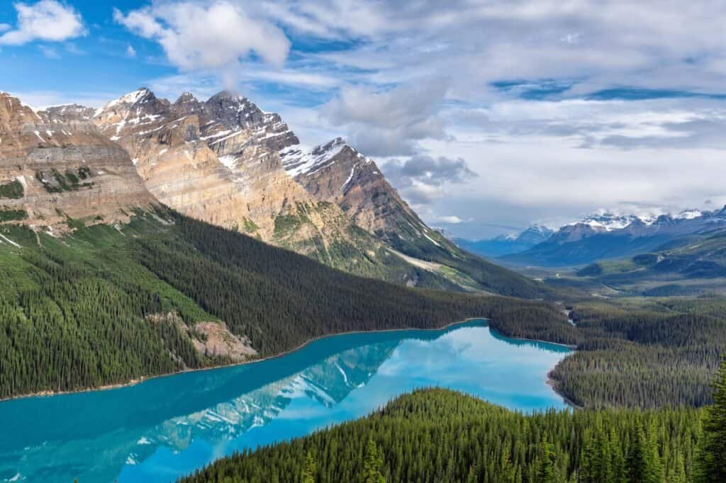 A scenic view of a turquoise lake surrounded by dense green forests and rugged snow-capped mountains under a partly cloudy sky, perfect for hiking and trekking tours.