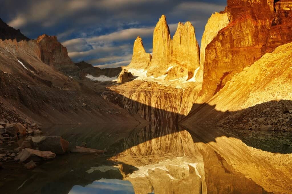 Dramatic mountain peaks reflect in a clear, tranquil lake under a sky filled with scattered clouds. The rocky landscape, perfect for hiking and trekking tours, is illuminated by warm, golden sunlight.