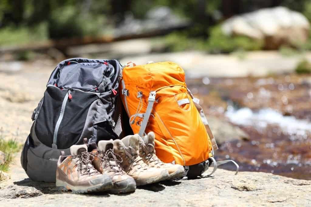 Two backpacks and two pairs of hiking boots rest on a rocky surface, waiting for their next adventure. In the background, a blurred stream meanders through lush greenery—a perfect scene for those who love hiking and trekking tours.