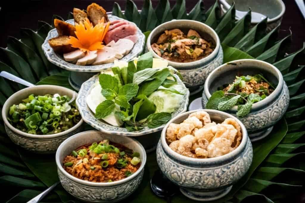 Assorted Thai dishes served in decorative bowls on a bed of green leaves, perfect for fueling your next hiking adventure, featuring vegetables, meats, and crispy pork rinds.