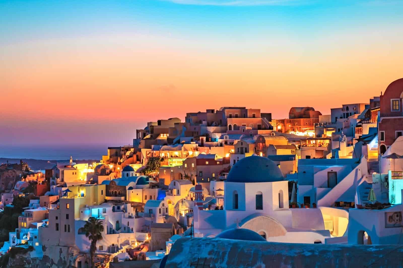 A vibrant sunset over a coastal town with colorful buildings, including a blue-domed white church, sets the scene for luxury adventure travel as it overlooks the sea.