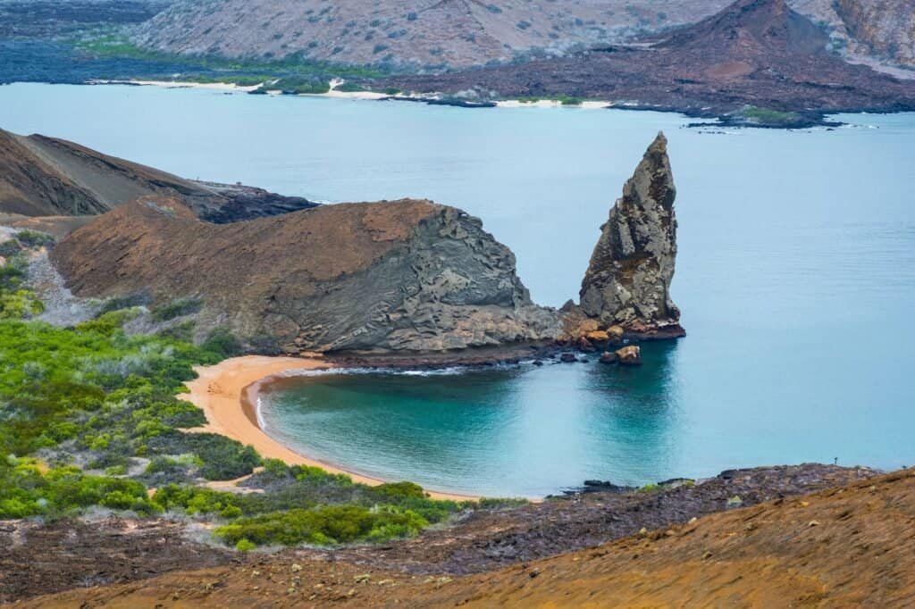 A rugged, triangular rock formation rises from a turquoise bay, embodying the essence of luxury adventure travel. The scene is framed by volcanic terrain and lush green shrubbery under a cloudy sky, promising an unforgettable escape into nature's grandeur.