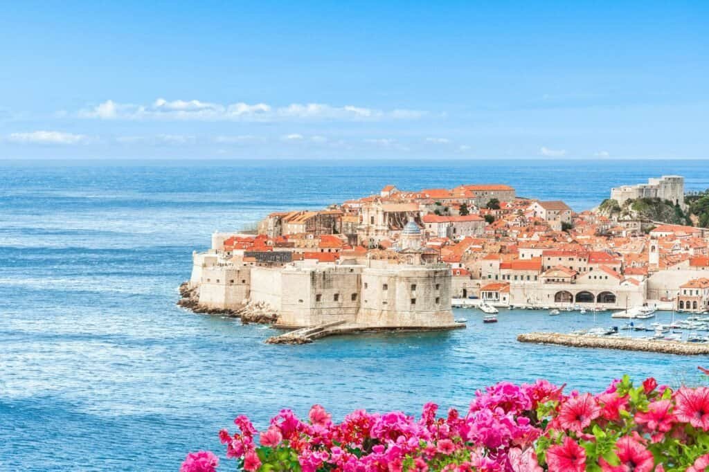 Experience the luxury of adventure travel in this historic coastal city, where orange rooftops and stone buildings meet the sea, and vibrant pink flowers bloom in the foreground.