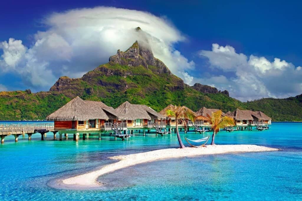 Overwater bungalows and palm trees on a small sandy island offer a luxury escape in turquoise waters, with a lush mountain and clouds painting an adventurous backdrop.
