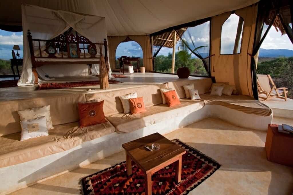 Experience luxury adventure travel in a beautifully designed tent featuring beige seating, patterned rugs, and a raised bed. Wooden furniture complements the large windows that invite the natural surroundings inside, creating a serene and lavish retreat amidst nature.