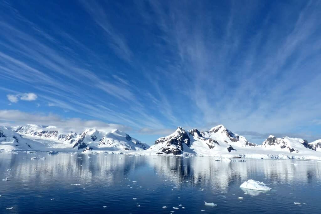 Snow-covered mountains reflected in a calm, icy body of water under a clear blue sky with wispy clouds provide the perfect backdrop for a luxury adventure.
