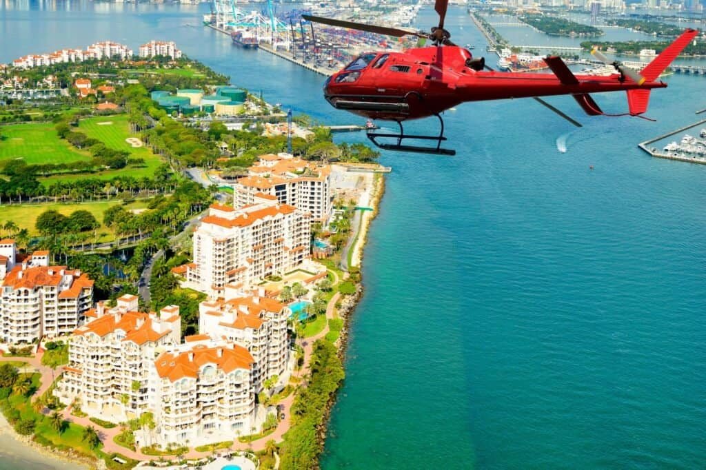 A red helicopter flies over a coastal city with green spaces, residential buildings, and a marina in view.