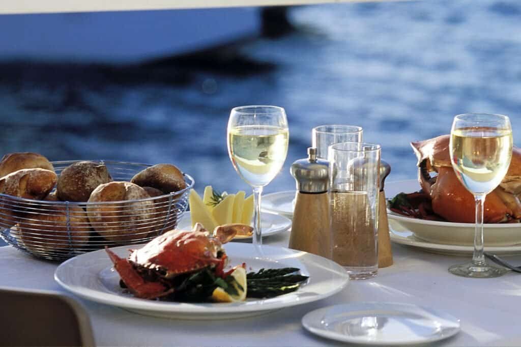 Table with two plates of seafood, two glasses of white wine, a basket of bread rolls, and condiments, near a waterfront.