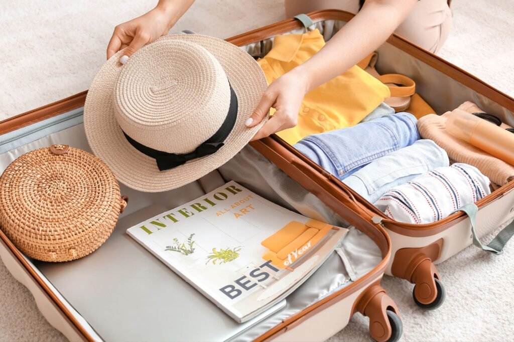 Person packing a suitcase with clothes, a straw hat, a round woven bag, and an interior design magazine.