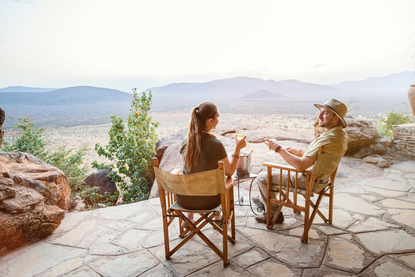 Two people sitting in chairs on a stone patio, enjoying drinks and a scenic view of mountains and valleys.