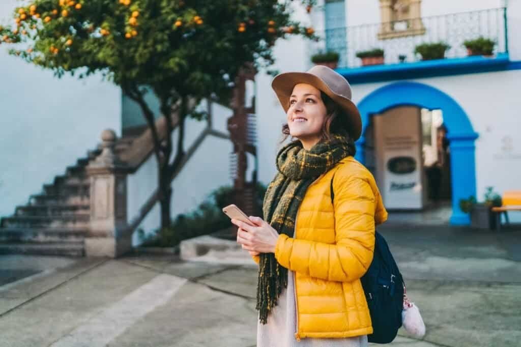 A person in a yellow jacket and hat stands outdoors, holding a phone, with a white building and orange tree nearby.