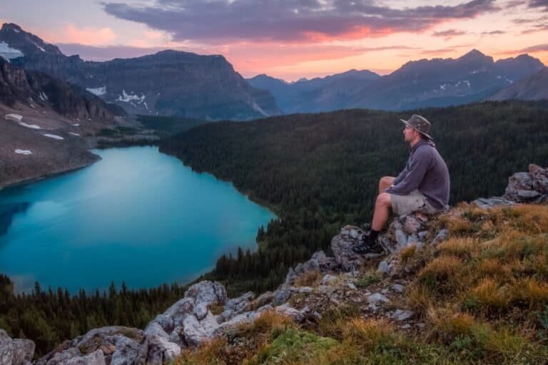 Person sitting on a rocky hill overlooking a turquoise lake surrounded by mountains at sunset.