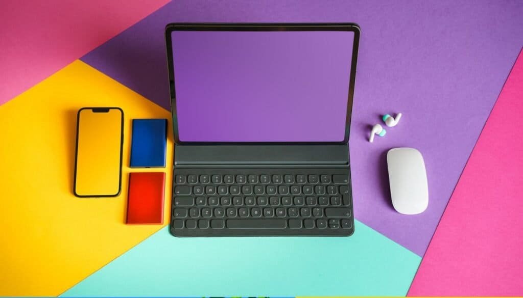 A vibrant, colorful background sets the stage for travel technology essentials: a tablet with keyboard, smartphone, colored cards, wireless earbuds, and mouse, all ready for your next adventure.