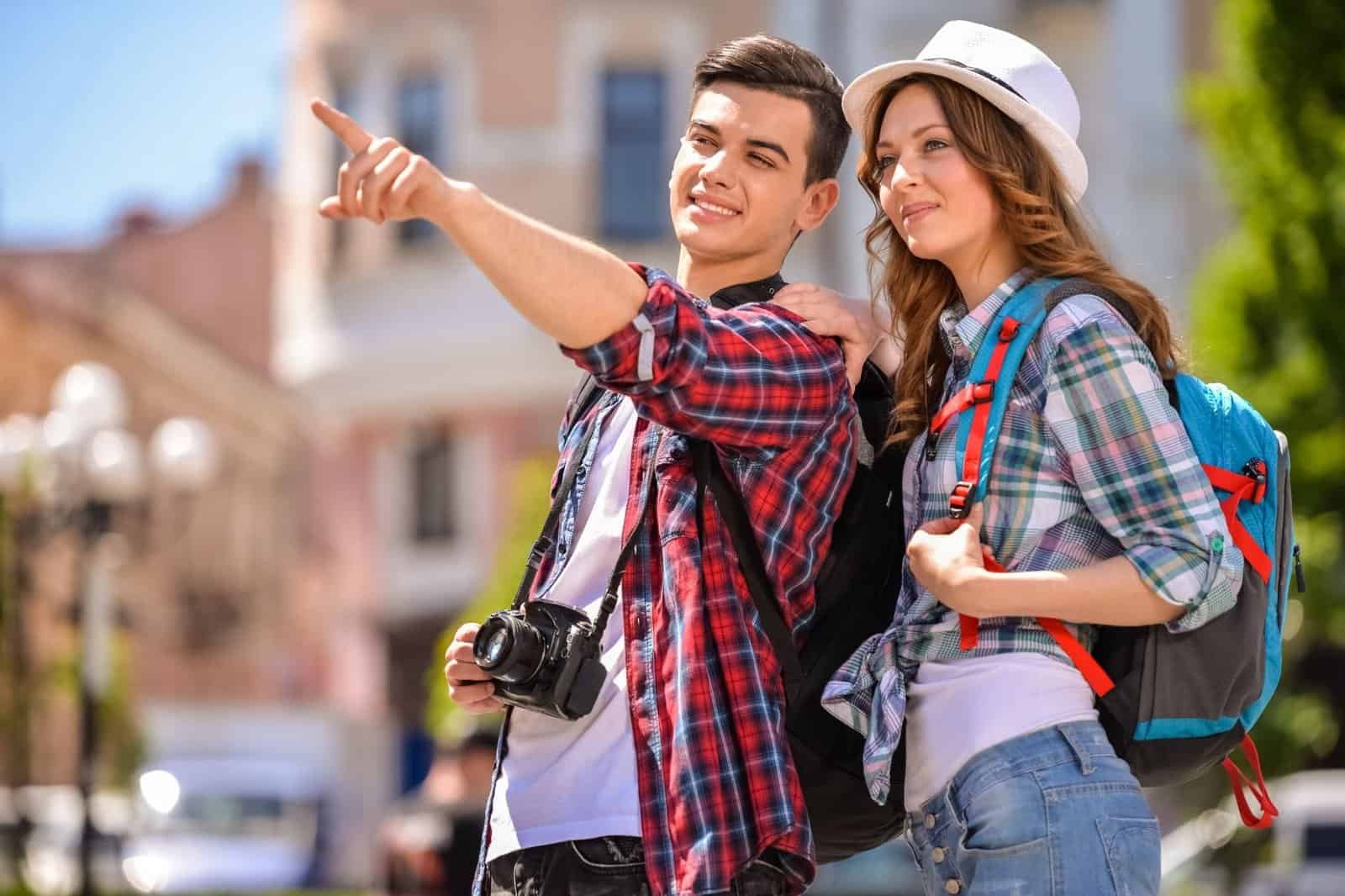 Two people in casual attire, wearing backpacks, smile while one points ahead. They stand outdoors in an urban setting, with trees and buildings in the background.