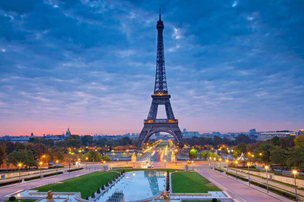 The Eiffel Tower at dusk, featured in many destination guides, stands majestic against a partly cloudy sky, surrounded by trees and paved pathways in Paris.