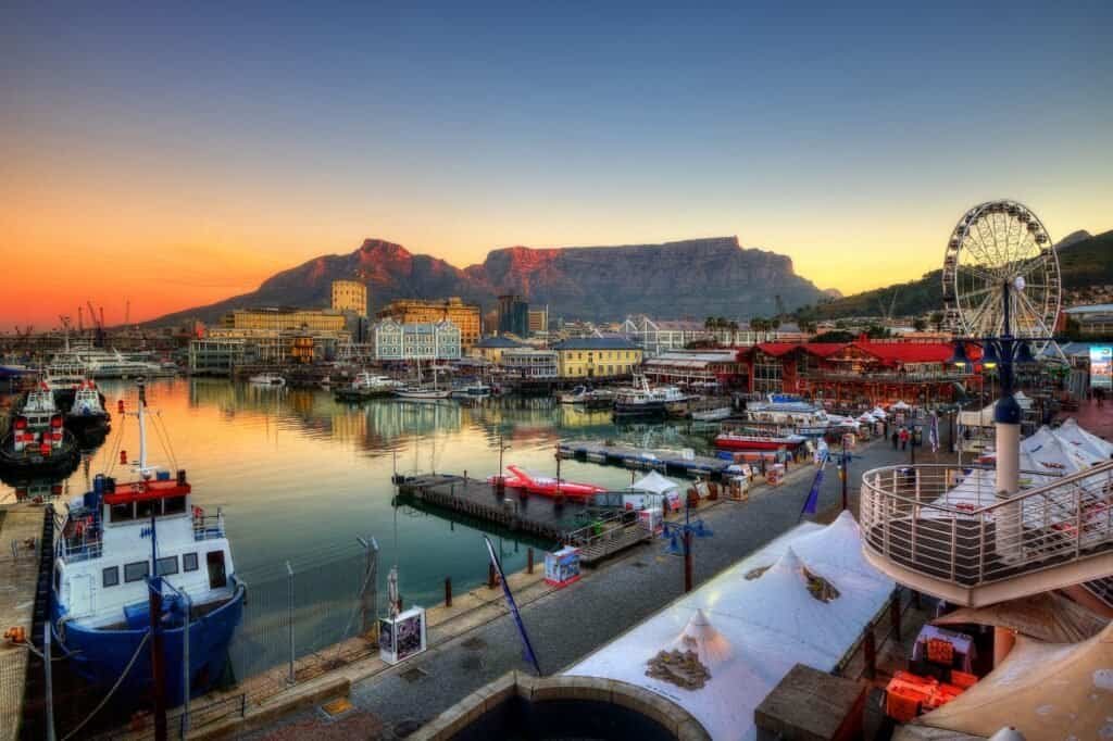 Experience the vibrant scene of a bustling harbor, where boats are docked beneath a colorful sky. A ferris wheel spins joyfully with majestic mountains as its backdrop, making this picturesque location a perfect highlight in any destination guide.