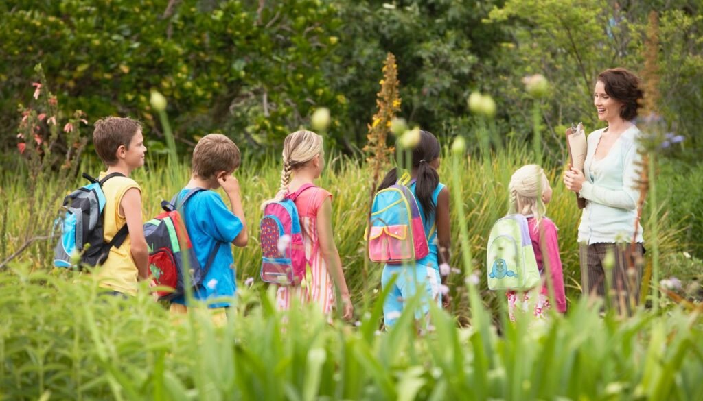 A teacher leads a group of children with colorful backpacks through a lush, green garden.