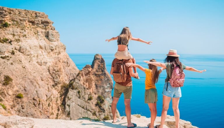 A family enjoys a scenic ocean view from a cliff, with one child perched happily on an adult's shoulders, experiencing the joy of family travel