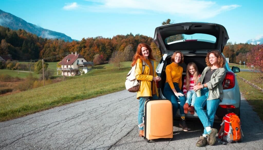 Four people with luggage pose by a car parked on a rural road, enjoying the scenic backdrop of hills and houses, capturing the essence of family travel.