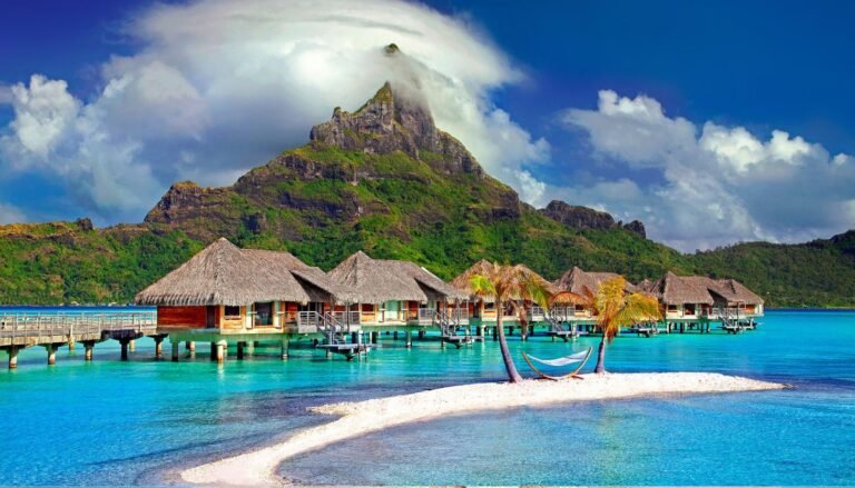 With luxury travel at its finest, bungalows over water with thatched roofs are set against a mountain backdrop. A small sandy island with palm trees and a hammock lies in the foreground, while clouds gently cover the mountain peak.