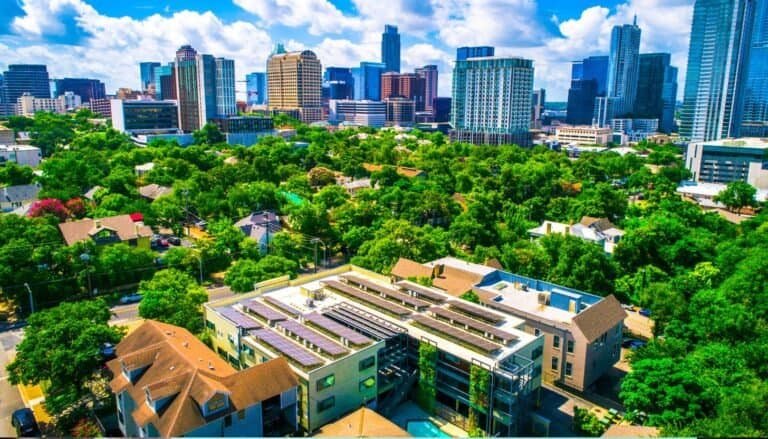 Aerial view of a cityscape with modern buildings, lush green trees, and a few residential houses in the foreground under a partly cloudy sky, perfectly capturing the essence of sustainable travel