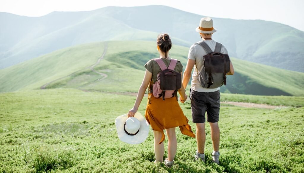Two people with backpacks stand on a grassy hill, embracing eco travel as they face a distant mountain range under a clear sky.