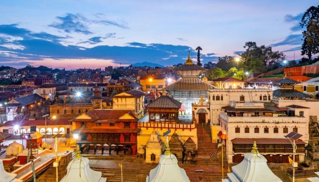 Colorful view of a temple complex, perfect for budget travel enthusiasts, with ornate buildings and bright lights at dusk, set against a backdrop of a city skyline and cloudy sky.