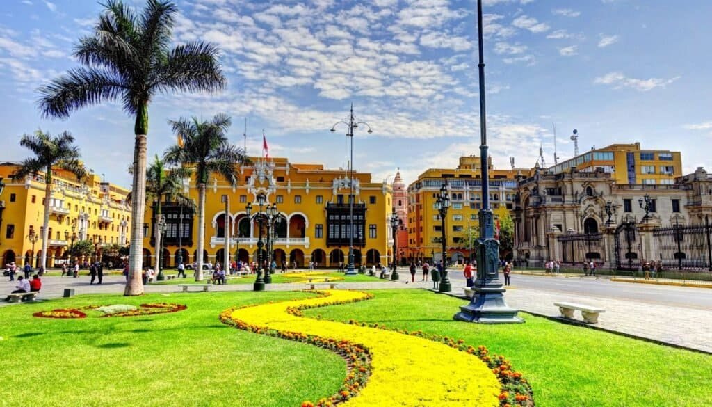 A vibrant city square with yellow buildings, palm trees, and a winding flower bed on green grass under a partly cloudy sky. Perfect for budget travel enthusiasts, people are walking and relaxing, soaking in the atmosphere without spending much.