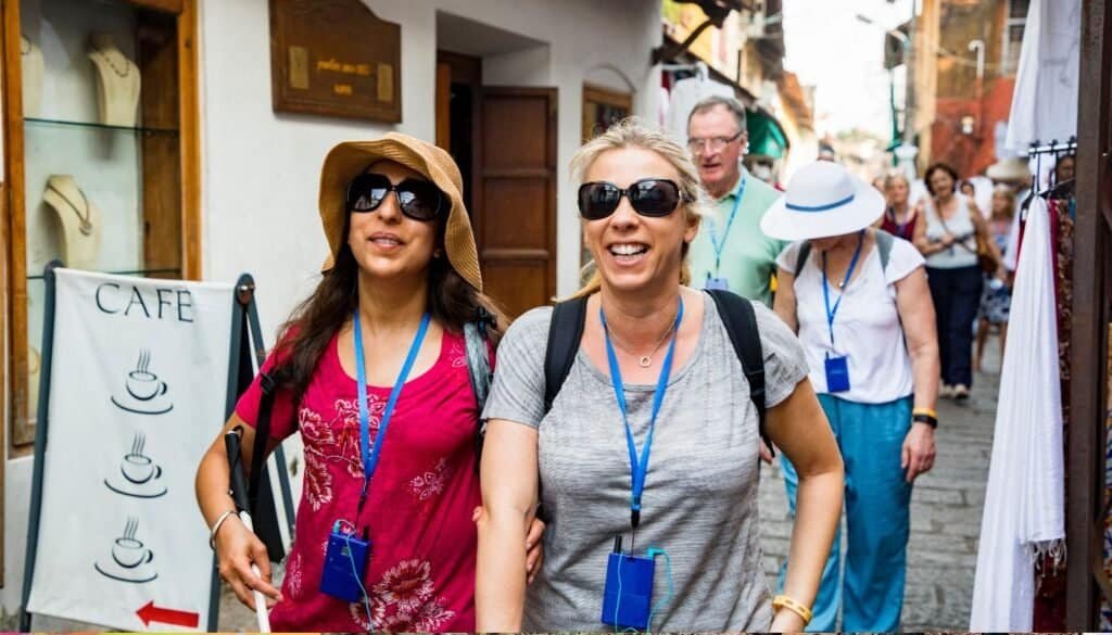 Two women walking arm in arm on a sunny street, wearing sunglasses and hats, with other people and shops in the background.