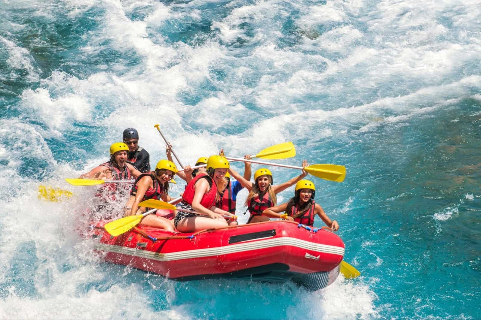 A group of people wearing helmets and life vests is white water rafting in a red inflatable boat on a turbulent river.