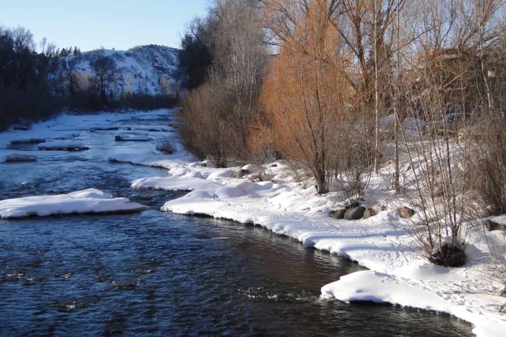 Winter river scene with snow-covered banks and leafless trees under a clear blue sky.