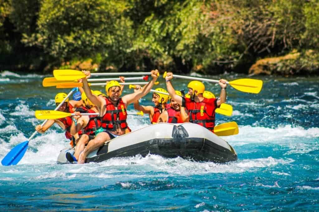 A group of people in helmets and life jackets are rafting on a river, raising their paddles with excitement.