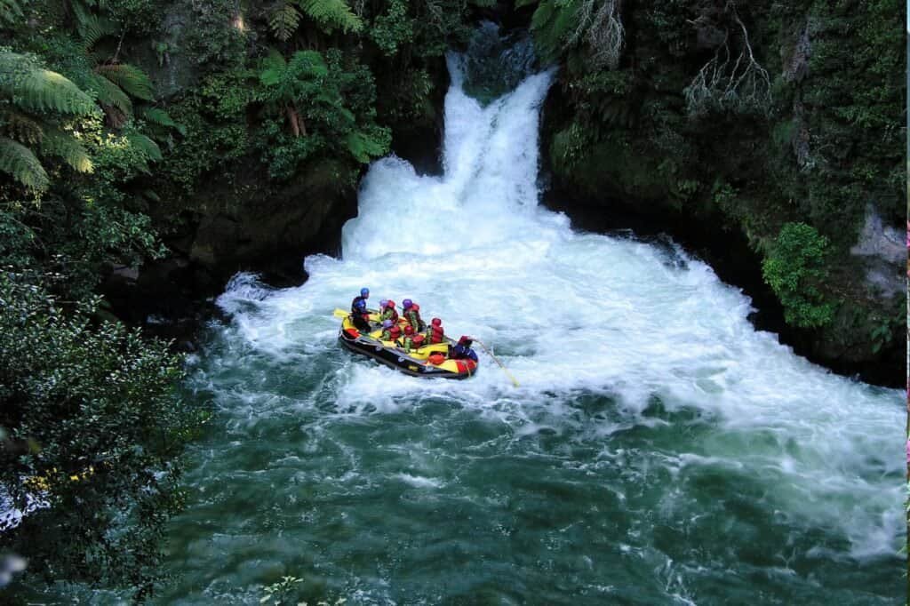 A group of people in helmets and life jackets are white-water rafting on a turbulent river near a small waterfall, surrounded by lush greenery.