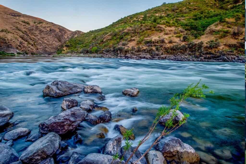 A river flows through a rocky landscape with hills in the background. The water appears rapid and clear, surrounded by greenery.