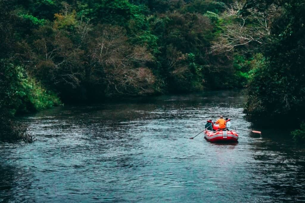 Two people in a red inflatable raft paddle down a calm river surrounded by dense green trees.