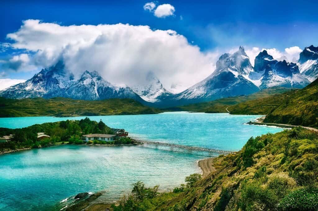 Mountain landscape with a turquoise lake, snow-capped peaks in the background, and a small building on the shore connected by a bridge.