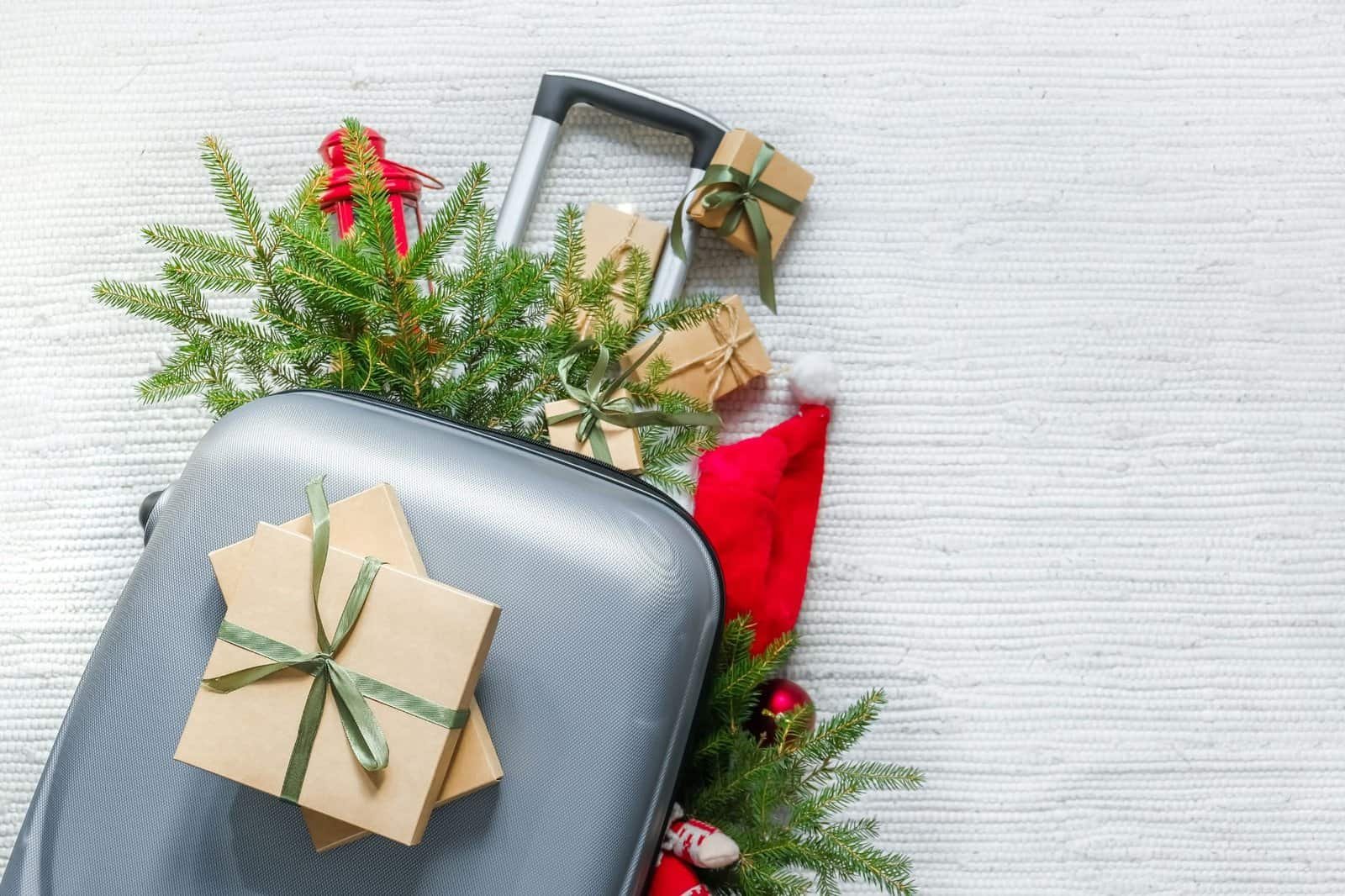 A gray suitcase is placed on a white carpet, surrounded by pine branches, wrapped gifts, and a Santa hat.