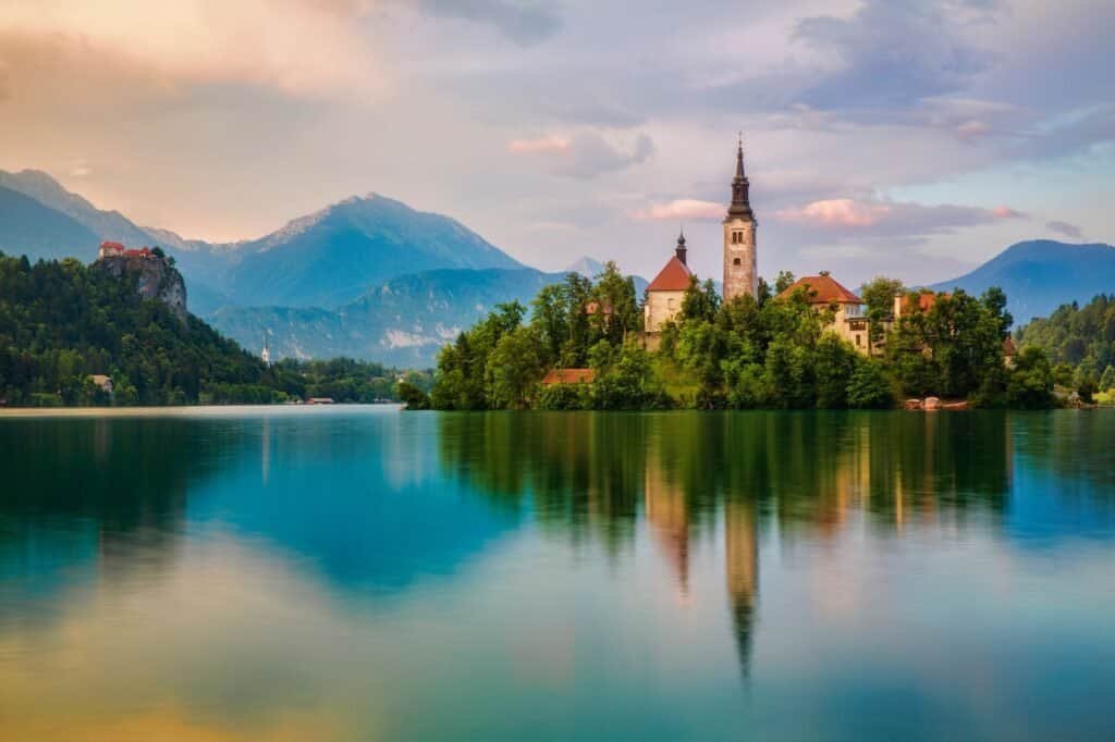 A serene lake with a small island featuring a church and surrounding trees, set against a backdrop of mountains under a partly cloudy sky, creates the perfect setting for a fall adventure.