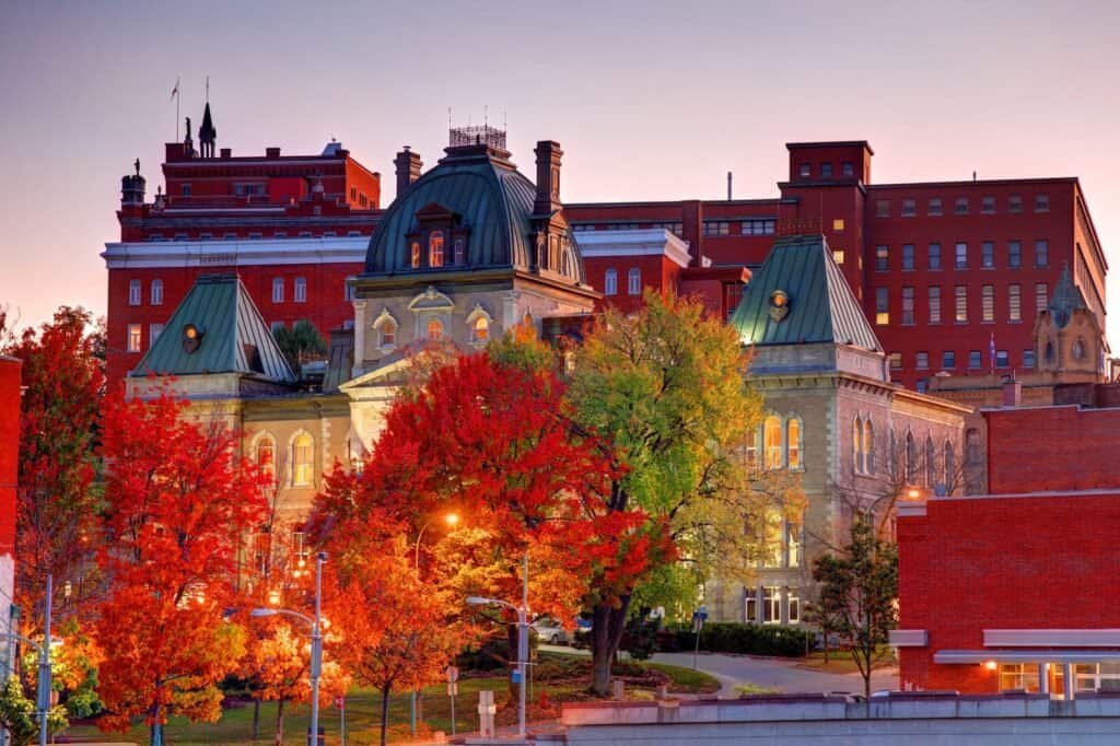 A historic building is surrounded by vibrant autumn trees, setting the scene for a perfect fall adventure, with a backdrop of red-brick structures glowing in the sunset.