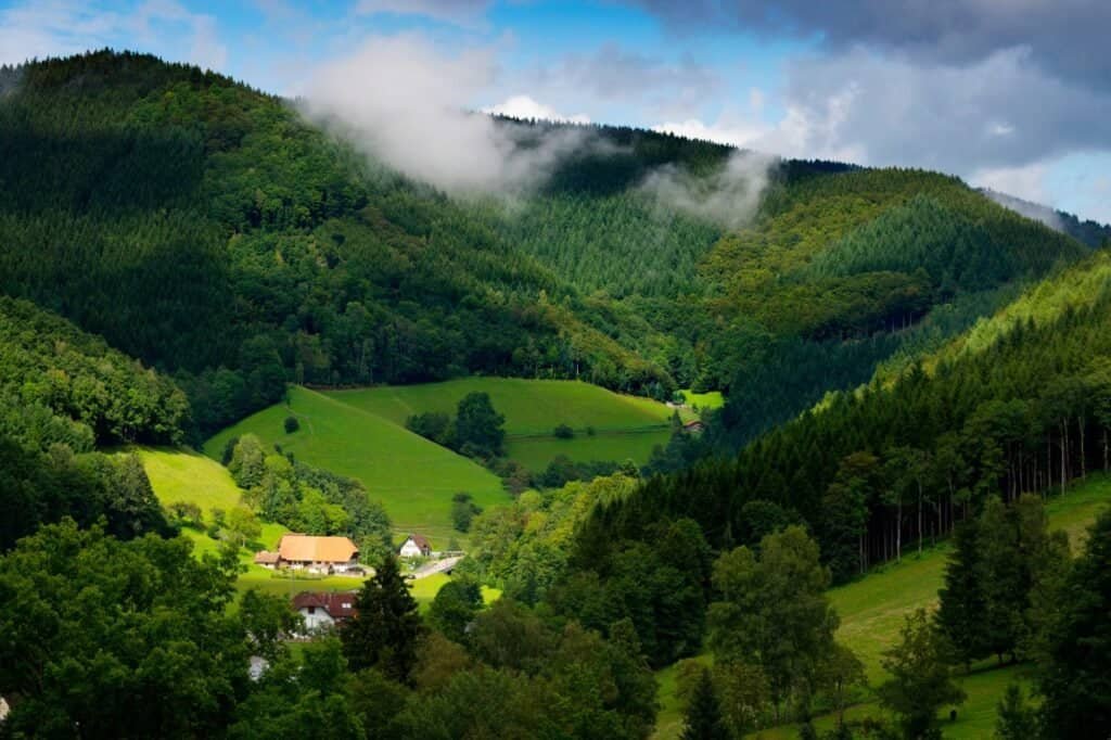 Aerial view of a lush green valley with a small village surrounded by dense forests, perfect for fall adventure travel. Rolling hills under a partly cloudy sky invite explorers to embrace the beauty of the season.