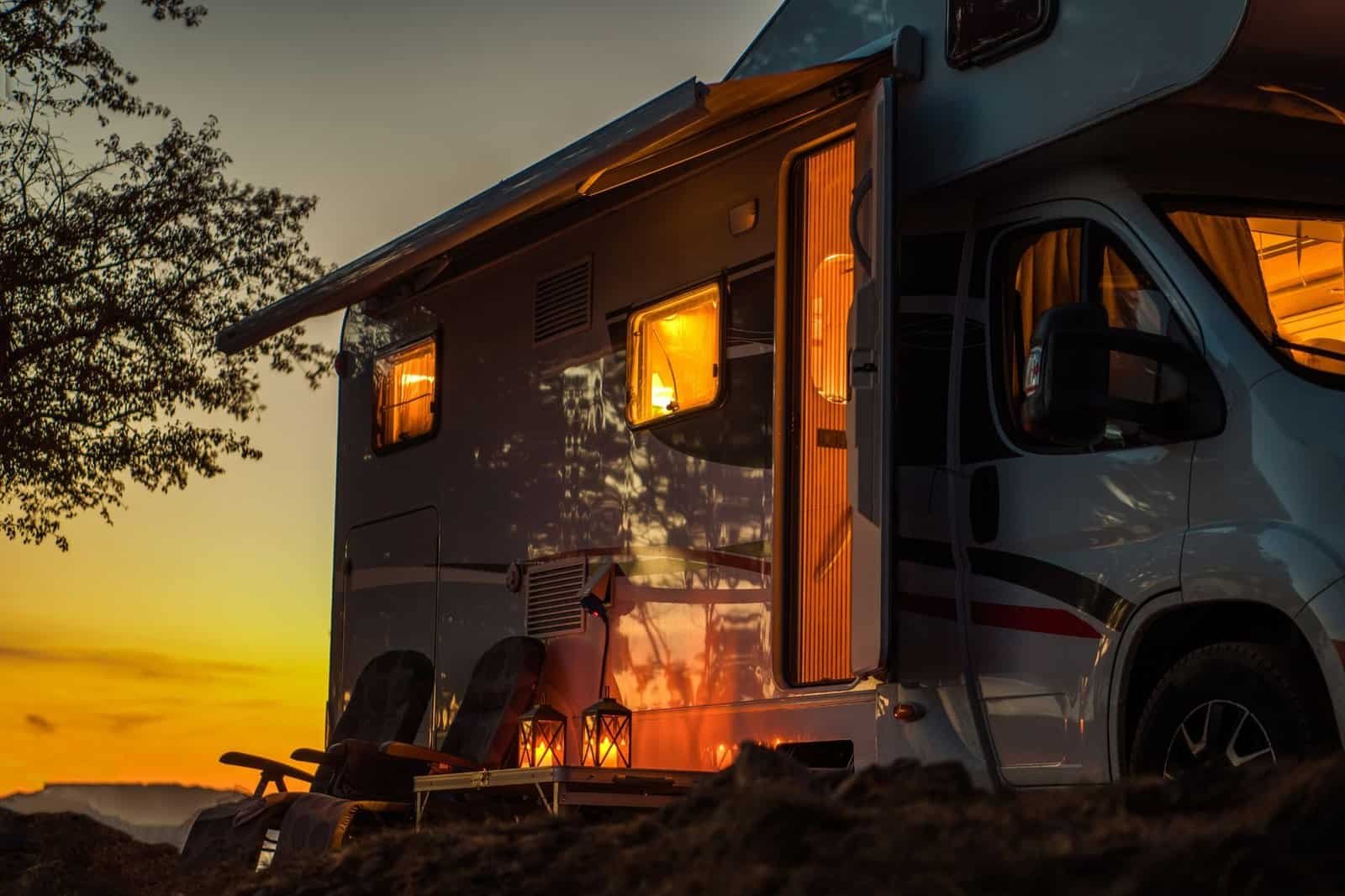 A camper van parked at sunset with its door open, warm interior lighting visible. Two chairs and a lantern are set outside under an extended awning, perfect for a cozy fall adventure travel experience.