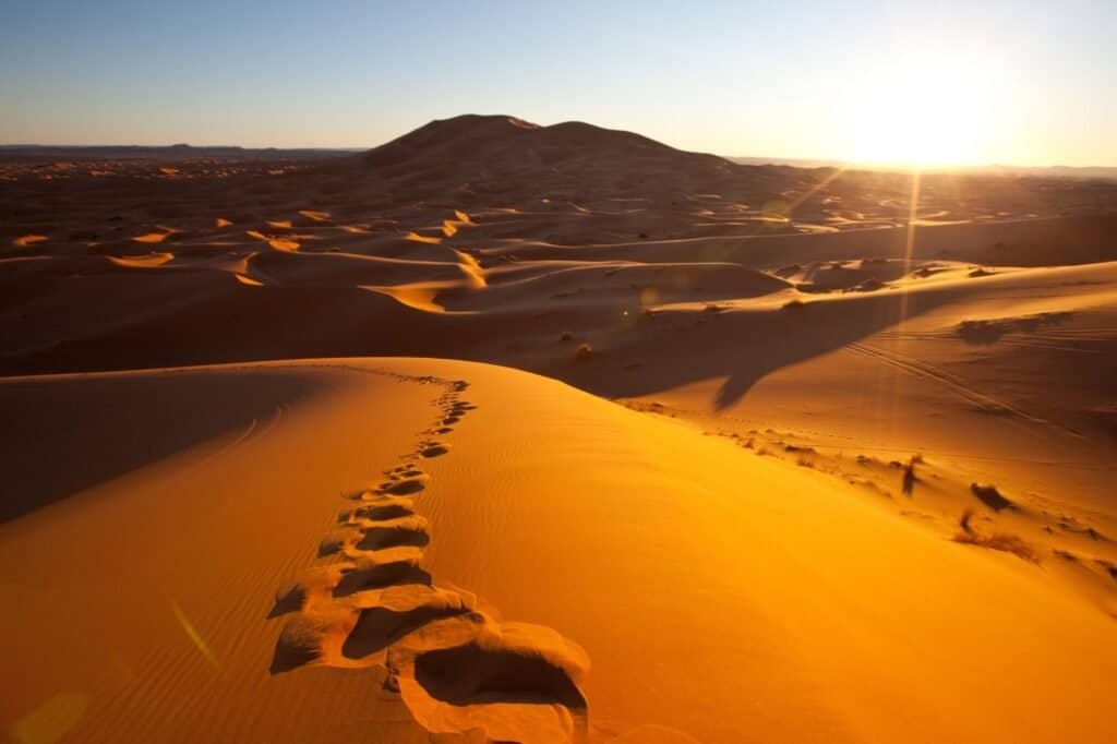 Footprints trail across sand dunes towards a distant hill, offering a serene backdrop for summer adventure ideas, as the sun sets in the desert landscape.